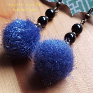 May include: A pair of earrings with blue fuzzy pom poms and black beads hanging from gold hooks. The earrings are labeled "Tapas for Two Gifts by Rosa Casado".