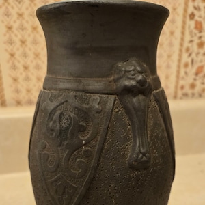 May include: A black ceramic vase with a textured surface and a raised design of a lion's head on the side. The vase has a wide mouth and a narrow base.