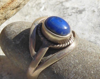 Può includere: Un anello d'argento con una pietra preziosa di lapislazzuli blu incastonata in una montatura a castone. L'anello ha un design a fascia intrecciata.