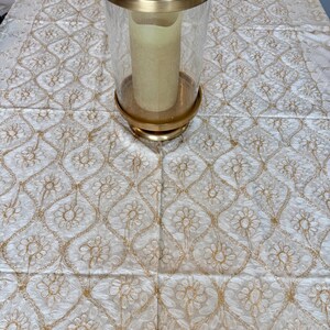 May include: A gold-colored metal and glass candle holder with a cream-colored candle inside. The candle holder sits on a white tablecloth with a gold floral embroidered pattern.