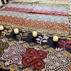 May include: A decorative table runner with intricate embroidered patterns in shades of gold, red, purple, and green. The runner is placed on a dark surface and adorned with small white candles. The design features floral and paisley motifs.