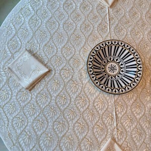 Könnte beinhalten: Eine cremefarbene Tischdecke mit einem gestickten Blumenmuster, zusammen mit passenden Servietten. Ein Teller mit schwarz-weißem Muster liegt auf der Tischdecke und erzeugt einen Kontrast in Texturen und Farben. Die Gesamtästhetik ist elegant und für das Essen geeignet.