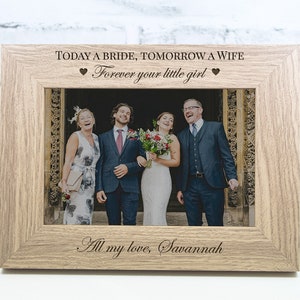 May include: A wooden picture frame with a photo of a bride and groom with their families. The frame has the text "TODAY A BRIDE, TOMORROW A WIFE" and "Forever your little girl" with hearts on either side. The bottom of the frame has the text "All my love, Savannah".
