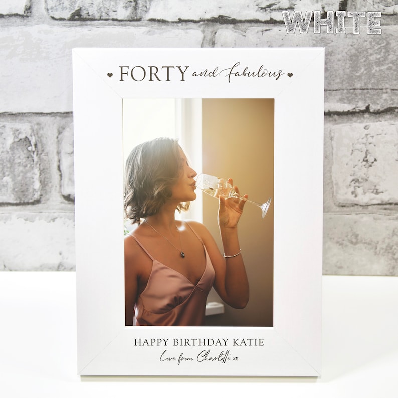Op de afbeelding: Wit fotolijstje met de tekst "FORTY and fabulous" bovenaan. Het lijstje bevat een foto van een persoon die een glas champagne vasthoudt. De tekst "HAPPY BIRTHDAY KATIE Love from Charlotte xx" staat onderaan het lijstje.