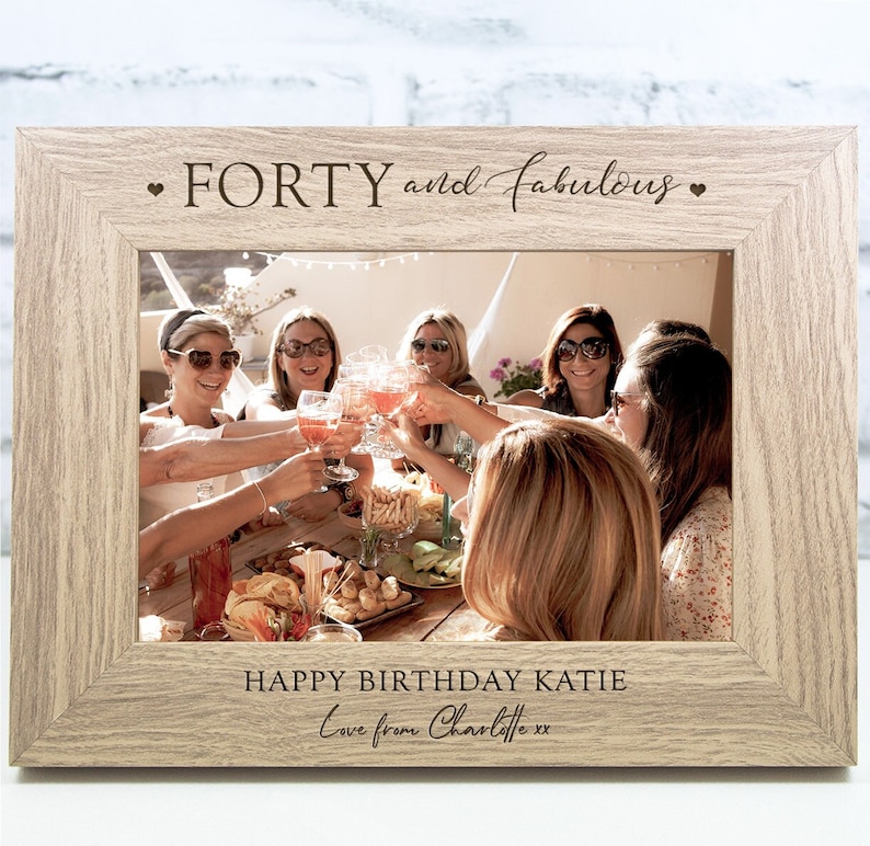 Op de afbeelding: Een licht houten fotolijst met een gepersonaliseerd bericht dat "FORTY and fabulous" en "HAPPY BIRTHDAY KATIE Love from Charlotte xx" luidt. De lijst bevat een foto van een groep vrienden die met drankjes vieren.