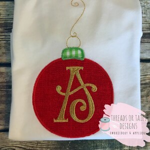 Op de afbeelding: Een wit T-shirt met een rood en gouden geborduurd kerstversiering. De versiering is rond met een groen-wit geruite strik aan de bovenkant. De letter "A" is in goud geborduurd in de versiering. De tekst "THREADS OR TAILS DESIGNS EMBROIDERY & APPLIQUE" is in zwart geborduurd in de rechterbenedenhoek van het shirt.
