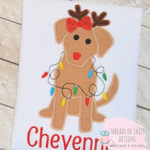 May include: Embroidered design of a brown dog wearing a red bow and reindeer antlers, holding a string of colorful Christmas lights. The name "Cheyenne" is embroidered in red below the dog. The design is on a white t-shirt. Threads or Tails Designs Embroidery & Applique is printed below the name.