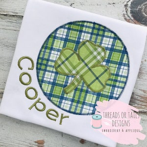 Peut inclure: Un t-shirt blanc avec une broderie verte d'un trèfle dans un cercle à carreaux bleus et verts. Le nom "Cooper" est brodé en vert sous le cercle. Le texte "Threads or Tails Designs Embroidery & Applique" est brodé en rose et vert dans le coin inférieur droit.