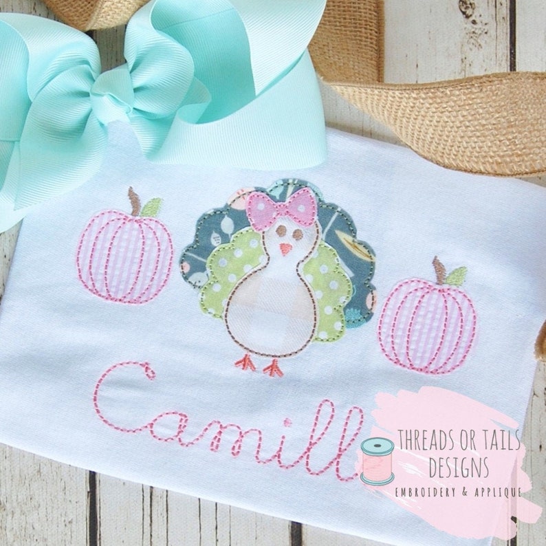 Puede incluir: Un pelele blanco con un pavo bordado en rosa y verde y dos calabazas rosas. El pelele est&aacute; personalizado con el nombre "Camille" en hilo rosa. El texto "Threads or Tails Designs Embroidery & Applique" tambi&eacute;n est&aacute; bordado en el pelele.