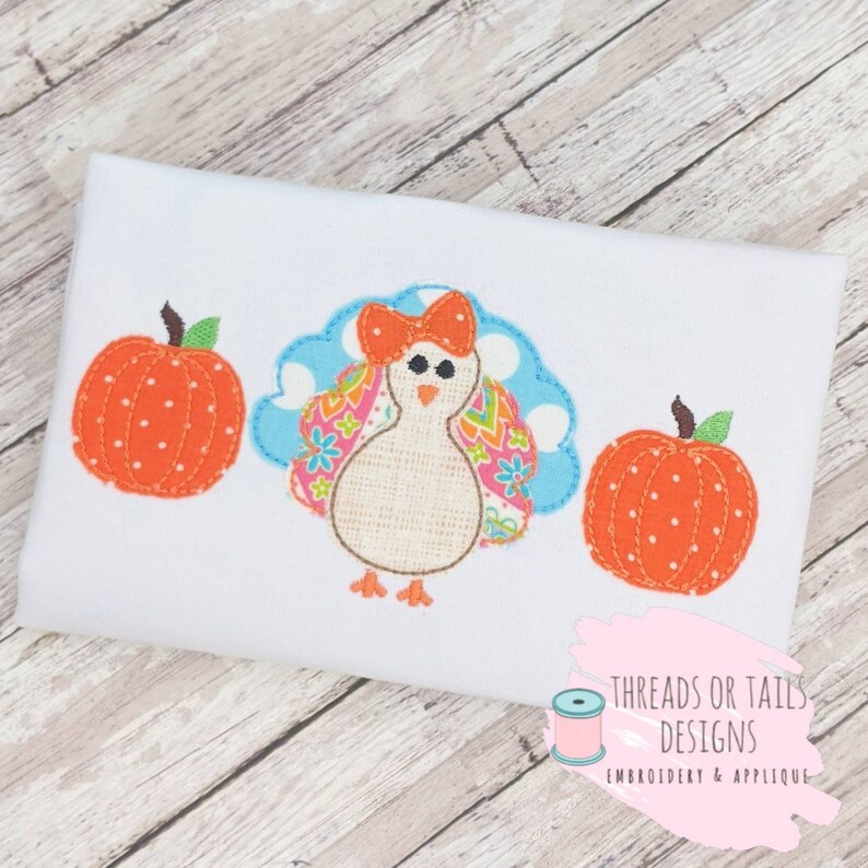 Puede incluir: Dise&ntilde;o bordado con un pavo con cola de estampado azul y blanco, cuerpo rosa y lazo rojo. El dise&ntilde;o est&aacute; flanqueado por dos calabazas naranjas con tallos verdes y lunares blancos.