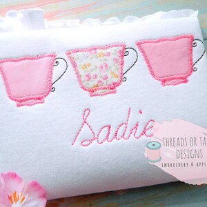 May include: White fabric with three pink teacups appliqued on it. The teacups have a pink outline and the middle teacup has a floral pattern. The name Sadie is embroidered in pink below the teacups. The text "Threads or Tails Designs Embroidery & Applique" is also embroidered in pink on the fabric.