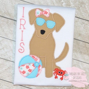 May include: Embroidered design of a brown dog wearing sunglasses and a pink bow, holding a blue and pink beach ball. A red crab is to the right of the dog. The text "IRIS" is stitched in pink thread on the left side of the design.