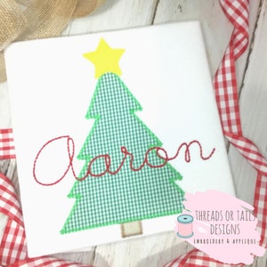 May include: White square with an embroidered Christmas tree design. The tree is green gingham with a yellow star on top. The name "Aaron" is embroidered in red script. The design is surrounded by red and white checkered ribbon.