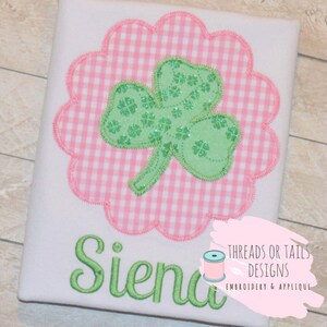 May include: A white t-shirt with a green shamrock applique on a pink and white gingham background. The name "Siena" is embroidered in green below the shamrock. The text "Threads or Tails Designs Embroidery & Applique" is in the bottom right corner.