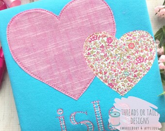 May include: A turquoise blue t-shirt with two pink hearts, one with a floral pattern. The word "isle" is embroidered in white on the shirt. The text "Threads or Tails Designs Embroidery & Applique" is written in white on a pink background.