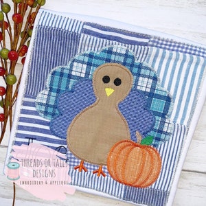 Könnte beinhalten: Ein brauner Truthahn mit blauen und weißen karierten Schwanzfedern und einem kleinen orangefarbenen Kürbis. Der Truthahn ist auf einem weißen Stoff mit blau-weißem Patchwork gestickt.