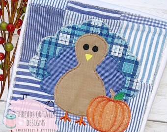 Op de afbeelding: Een bruine kalkoen met blauwe en witte geruite staartveren en een kleine oranje pompoen. De kalkoen is geborduurd op een witte stof met blauw-witte patchwork.
