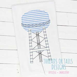 May include: A blue and white striped water tower applique embroidery design. The design features a water tower with a round top and a support structure. The text "THREADS OR TAILS DESIGNS APPLIQUE + EMBROIDERY" is at the bottom of the design.