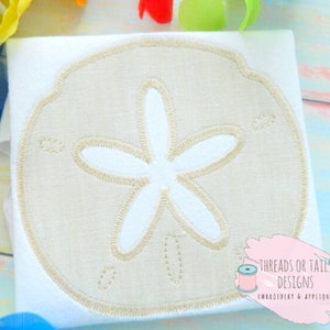 May include: A white t-shirt with a beige sand dollar embroidered on the front. The sand dollar has a white five-pointed star in the centre. The text "Threads or Tails Designs Embroidery & Applique" is visible in the bottom right corner of the image.