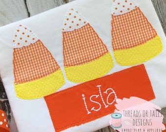 May include: A white t-shirt with three orange and yellow candy corn appliques. The shirt has an orange rectangle with the name "Isla" embroidered in white.