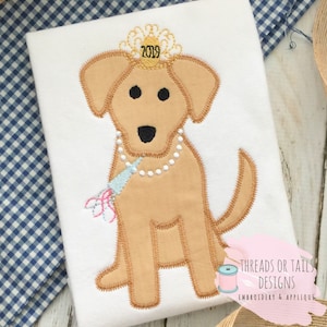 May include: Embroidered design of a brown dog wearing a gold crown with the year 2019 on it, a pearl necklace, and a blue and pink party horn. The design is on a white t-shirt. The text "Threads or Tails Designs Embroidery & Applique" is in the bottom right corner.