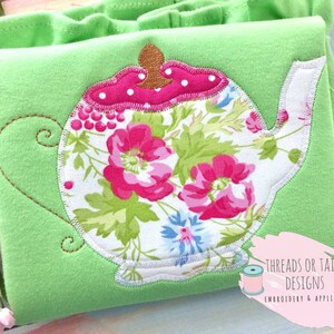 May include: A white teapot with a floral pattern and pink accents. The teapot is embroidered on a green fabric background. The text "Threads or Tails Designs Embroidery & Applique" is visible in the lower right corner.