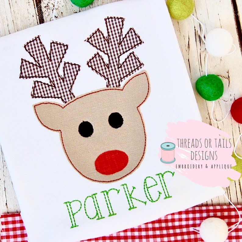 May include: A white onesie with a brown and white reindeer applique. The reindeer has a red nose and black eyes. The onesie has the name "Parker" embroidered in green on the front.