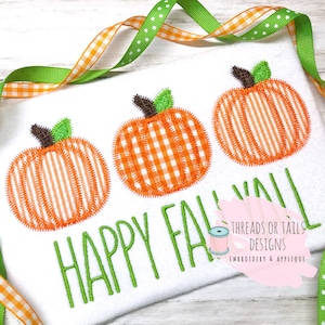 May include: Embroidered design featuring three orange pumpkins with a gingham pattern and green stems. The text "Happy Fall" is stitched in green below the pumpkins. The design is on a white fabric background.