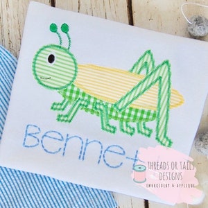 May include: A white onesie with a green and yellow embroidered grasshopper and the name "Bennett" stitched in blue thread. The onesie is decorated with a small embroidered spool of thread and the text "Threads or Tails Designs Embroidery & Applique".