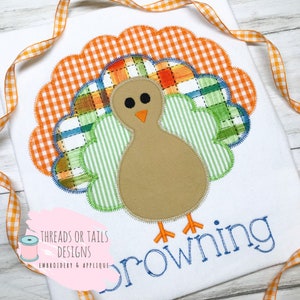Puede incluir: Una camiseta blanca con una aplicación bordada de un pavo marrón con plumas de cola de cuadros verdes y naranjas. El texto "Browning" está bordado en azul debajo del pavo. El texto "Threads or Tails Designs Embroidery & Applique" está bordado en negro en la esquina inferior izquierda.