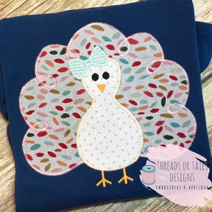 May include: A white turkey applique with a blue bow on a navy blue fabric. The turkey's tail feathers are a colorful pattern of red, green, and brown.