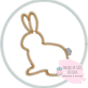 May include: A brown bunny silhouette embroidered in a circle with a light blue outline. The bunny is facing right and has a small gray tail. The text "Threads or Tails Designs Embroidery & Applique" is in the lower right corner of the image.
