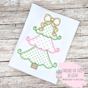 May include: A green and pink Christmas tree applique with a gold bow on top. The tree is decorated with white polka dots. The applique is on a white t-shirt.
