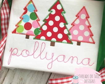 May include: A white shirt with three embroidered Christmas trees in red, pink, and green with white polka dots. The name "Pollyana" is embroidered in pink below the trees.