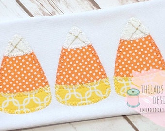 May include: Three embroidered candy corn appliques in orange, yellow, and white with a polka dot pattern on the orange section. The appliques are stitched on a white fabric background.