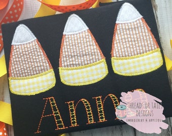 May include: Black fabric with three embroidered candy corn designs. Each candy corn has a white top, a brown and orange checkered center, and a yellow and white checkered bottom. The name "Ann" is embroidered below the candy corn.