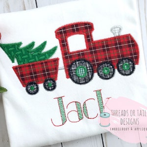 May include: White t-shirt featuring a red plaid train pulling a cart with a green Christmas tree. The train has black wheels with green centers. The name "Jack" is embroidered in green and red thread.