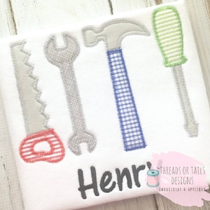 Peut inclure: Un t-shirt blanc avec une scie, une clé, un marteau et un tournevis gris brodés dessus. Le marteau a un manche à carreaux bleu et blanc. Le tournevis a un manche rayé vert et blanc. Le t-shirt porte également le nom "Henry" brodé en fil noir. Le coin inférieur droit du t-shirt porte le texte "Threads or Tails Designs Embroidery & Applique" en fil noir.