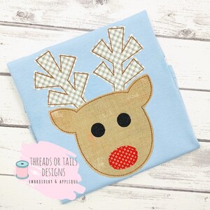 May include: A light blue fabric with a brown and red reindeer applique. The reindeer has a red nose with white polka dots, black eyes, and brown antlers with a white and gray checkered pattern. The fabric has the text "THREADS OR TAILS DESIGNS EMBROIDERY & APPLIQUE" in a pink box.