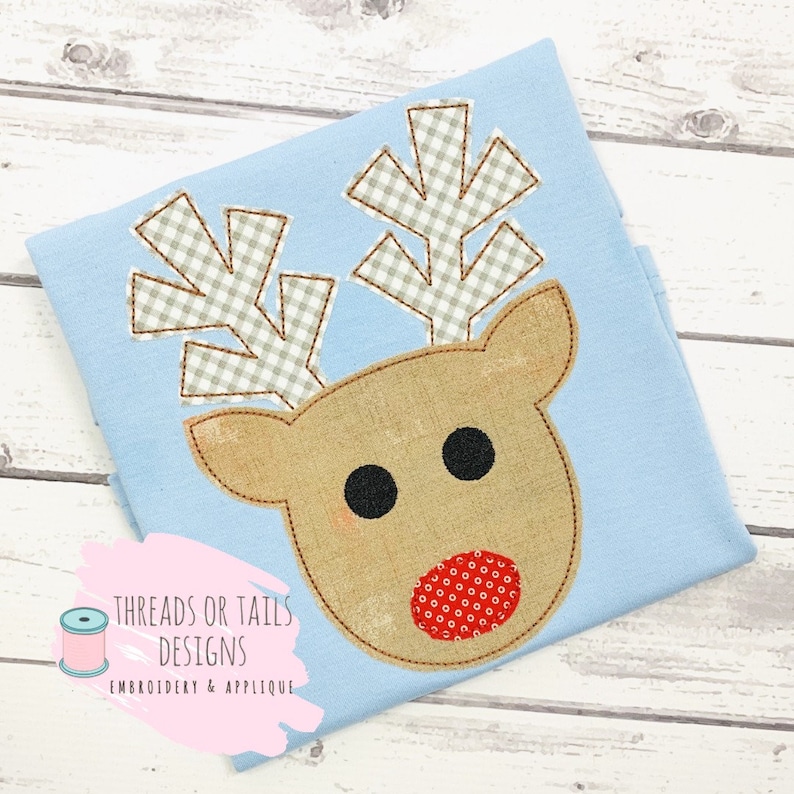 May include: A light blue fabric with a brown and red reindeer applique. The reindeer has a red nose with white polka dots, black eyes, and brown antlers with a white and gray checkered pattern. The fabric has the text "THREADS OR TAILS DESIGNS EMBROIDERY & APPLIQUE" in a pink box.