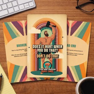 May include: Oracle cards with a retro design. The central card shows a muscular man with a hammer, and the text "DOES IT HURT WHEN YOU DO THAT? DON'T DO THAT." Other cards have text like "BROKEN" and "BAD END."