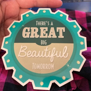 K&ouml;nnte beinhalten: Ein kreisf&ouml;rmiger Aufkleber in T&uuml;rkis und Creme mit dem Text "THERE'S A GREAT BIG BEAUTIFUL TOMORROW" in einer Schriftart im Vintage-Stil. Der Aufkleber hat einen gezackten Rand und wird von einer Hand gehalten.