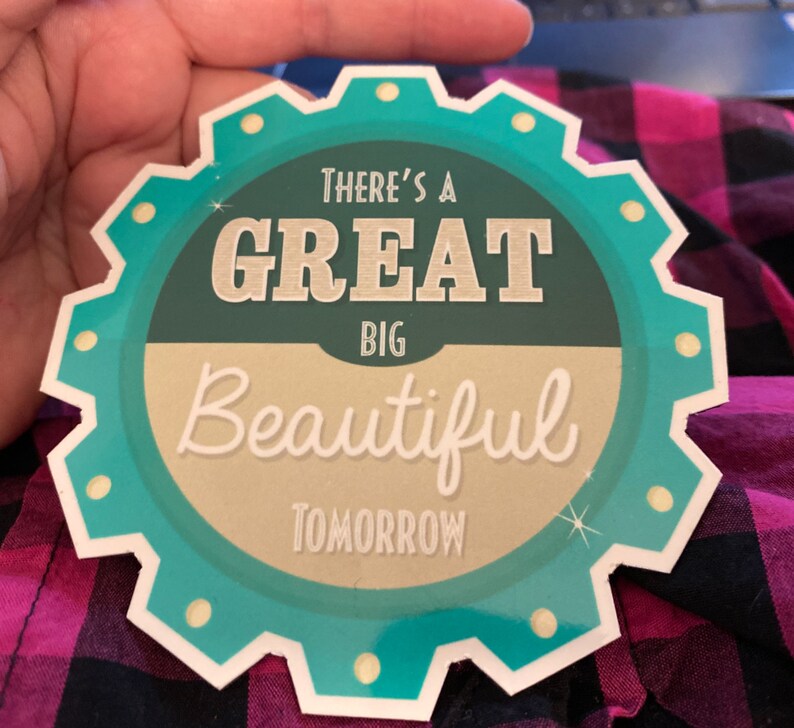 K&ouml;nnte beinhalten: Ein kreisf&ouml;rmiger Aufkleber in T&uuml;rkis und Creme mit dem Text "THERE'S A GREAT BIG BEAUTIFUL TOMORROW" in einer Schriftart im Vintage-Stil. Der Aufkleber hat einen gezackten Rand und wird von einer Hand gehalten.