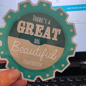 K&ouml;nnte beinhalten: Ein t&uuml;rkis-cremefarbener Aufkleber mit dem Text "THERE'S A GREAT BIG BEAUTIFUL TOMORROW" in einer stilisierten Schriftart. Der Aufkleber hat einen zahnradartigen Rand mit kleinen goldfarbenen Kreisen. Der Aufkleber wird vor einem Laptop gehalten.