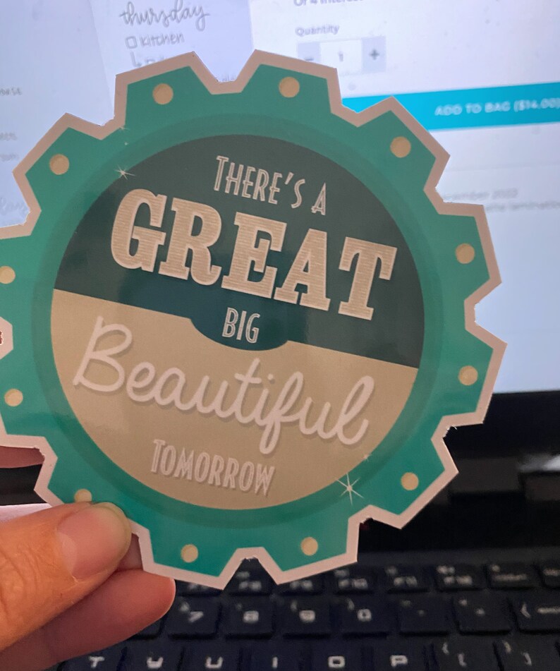 K&ouml;nnte beinhalten: Ein t&uuml;rkis-cremefarbener Aufkleber mit dem Text "THERE'S A GREAT BIG BEAUTIFUL TOMORROW" in einer stilisierten Schriftart. Der Aufkleber hat einen zahnradartigen Rand mit kleinen goldfarbenen Kreisen. Der Aufkleber wird vor einem Laptop gehalten.