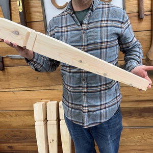 K&ouml;nnte beinhalten: Ein unfertiges M&ouml;belbein aus hellem Holz mit rechteckiger Oberseite und konischem Design. Das Bein hat ein geschnitztes Detail in der N&auml;he der Oberseite. Eine Person in einem karierten Hemd h&auml;lt das Bein. Im Hintergrund sind weitere &auml;hnliche Beine zu sehen.