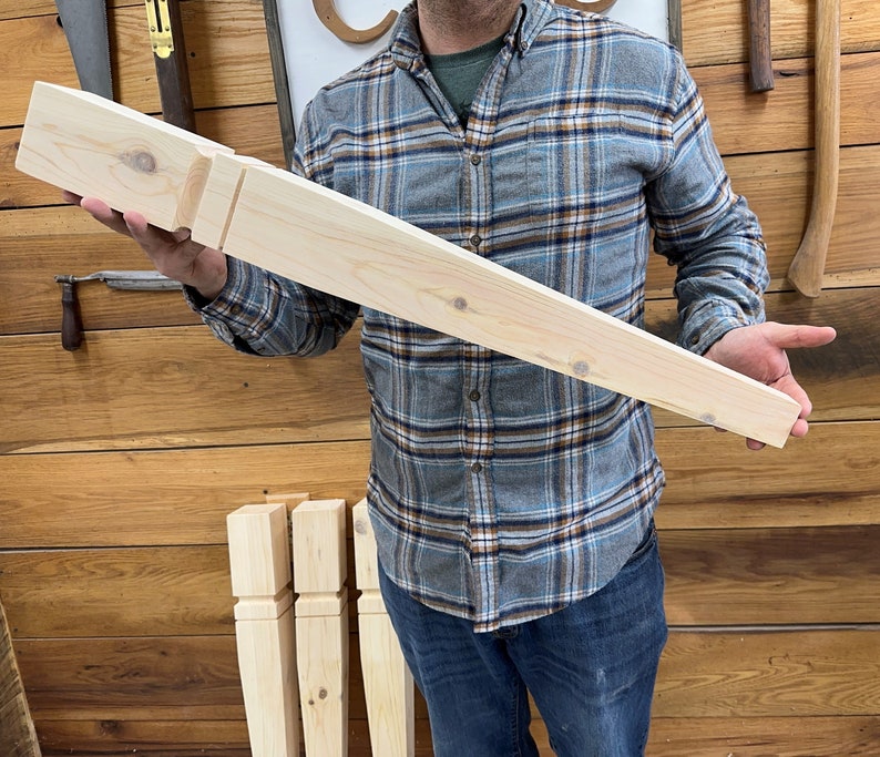 K&ouml;nnte beinhalten: Ein unfertiges M&ouml;belbein aus hellem Holz mit rechteckiger Oberseite und konischem Design. Das Bein hat ein geschnitztes Detail in der N&auml;he der Oberseite. Eine Person in einem karierten Hemd h&auml;lt das Bein. Im Hintergrund sind weitere &auml;hnliche Beine zu sehen.