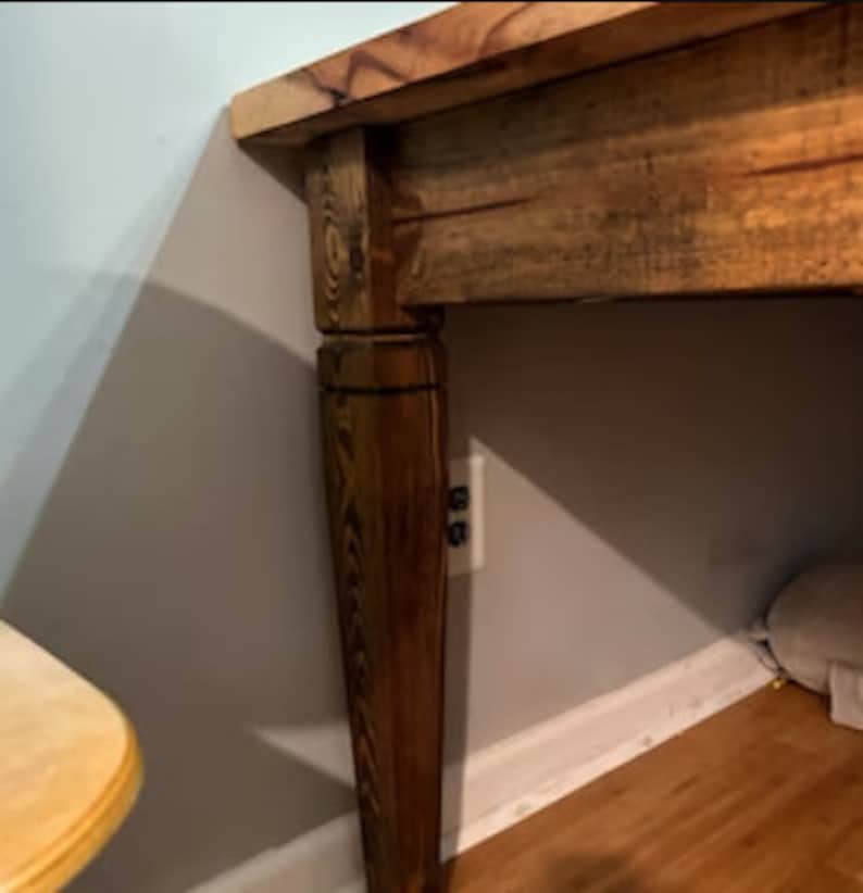K&ouml;nnte beinhalten: Nahaufnahme eines Holztisches mit dunkler Beize. Der Tisch hat eine dicke Platte und ein gedrechseltes Bein. Das Bein ist dunkelbraun und hat einen dekorativen Ring in der N&auml;he der Oberseite. Der Tisch steht vor einer hellblauen Wand.