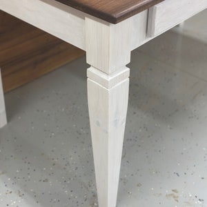 K&ouml;nnte beinhalten: Nahaufnahme eines Holztisches mit zweifarbigem Design. Die Tischplatte ist ein sattes, dunkles Braun, w&auml;hrend die Beine und die Zarge in einem hellen, cremewei&szlig;en Farbton lackiert sind. Die Tischbeine haben ein klassisches, leicht konisches Design.