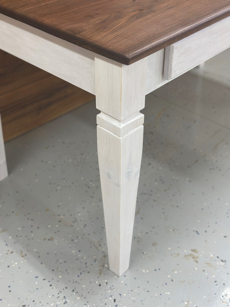 K&ouml;nnte beinhalten: Nahaufnahme eines Holztisches mit zweifarbigem Design. Die Tischplatte ist ein sattes, dunkles Braun, w&auml;hrend die Beine und die Zarge in einem hellen, cremewei&szlig;en Farbton lackiert sind. Die Tischbeine haben ein klassisches, leicht konisches Design.